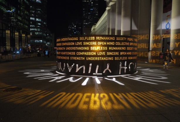 Humanity movement art installation in Sir John A Macdonald Plaza in front of Union Station, by Masai Ujiri. metal cylinder with words cut out, a light shines through from the middle and the words show as lights on the sidewalk
