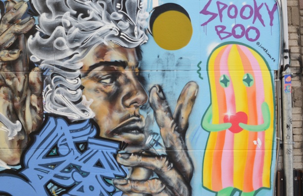 street art by Rowell Soller of black man's face in profile with calligraphy in white and blue as hair. Beside it is pink and yellow striped ghost figure called spooky boo by June Kim