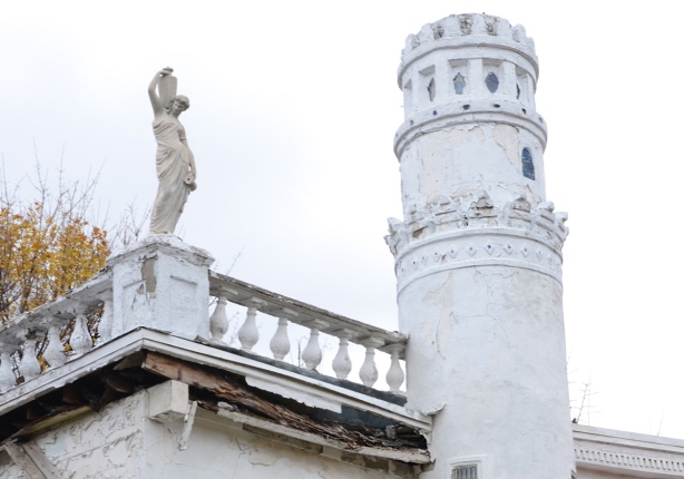 crooked turret beside railing around rooftop terrace with stone statue of a woman (greek goddess?) holding an urn 