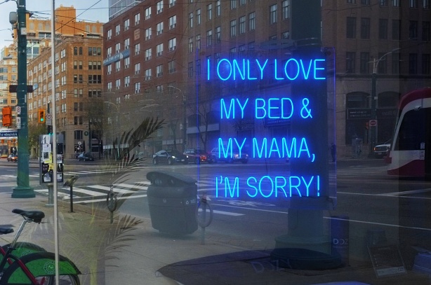 blue lights in a store window in the shape of letters and words that say I only love my bed and my mama, I'm sorry! also reflections of street scene in the window including TTC streetcar and intersection