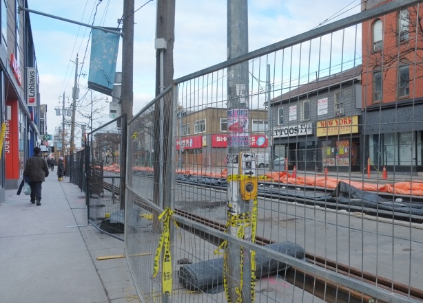woman walking on the sidewalk between store fronts and construction fence, TTC track work being done on Queen West 