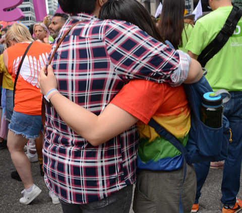 back view of two people hugging, one in a plaid shirt and the other in a rainbow t shirt