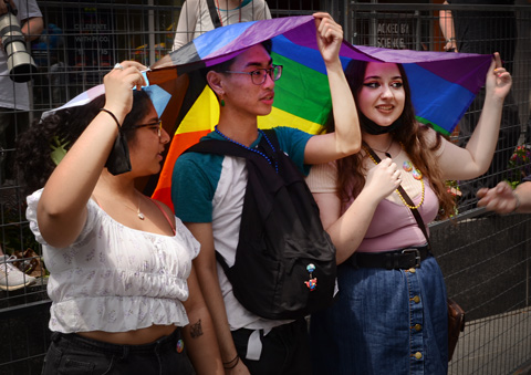 three young people holding a rainbow type flag over their heads as they watch pride parade