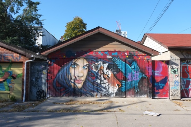painted garages in David French Lane, the one in the middle has a person with long black hair and blue eyes beside a tiger head in profile and a blue bird taking flight