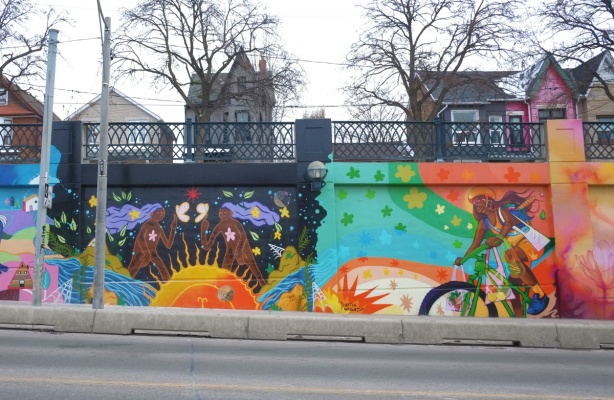 two murals on a concrete wall. on the right is a cyclist painted by Curtia Wright and on the left is a scene with two brown figures, a male and a female, standing above a yellow and orange sun