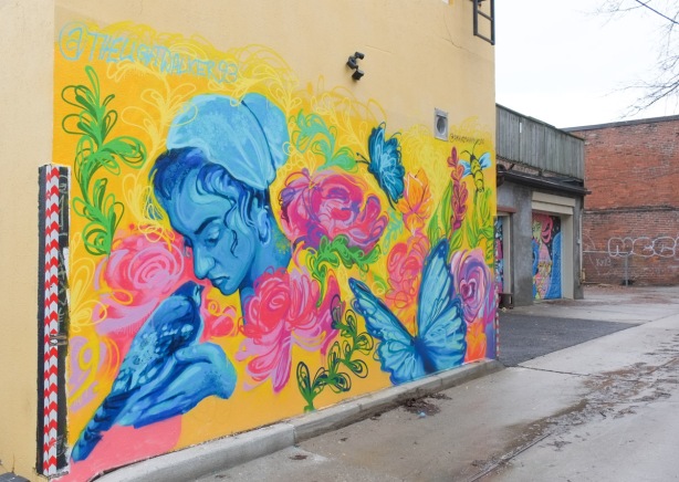 mural by Amani Muhammad, a woman in blue holds a blue pigeon, pink and red flowers around them as well as blue butterflies