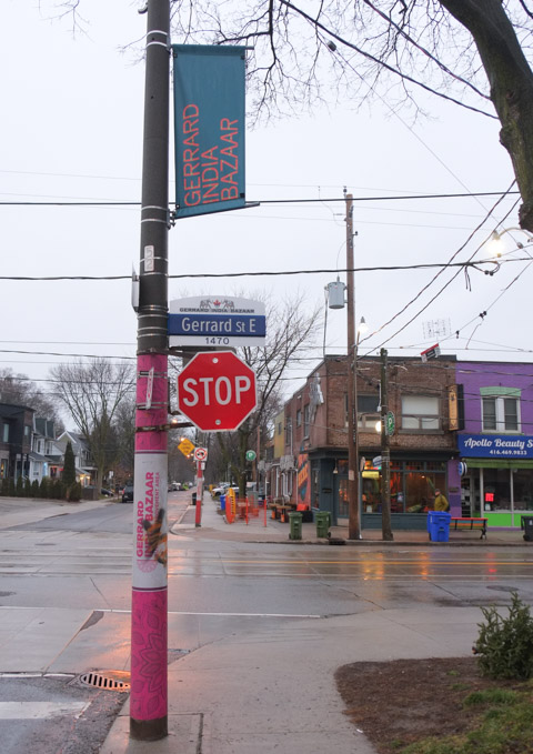 utility pole with stop sign on Gerrard, pink pole with gerrard india bazaar banner on the top, stores on Gerrard in the background