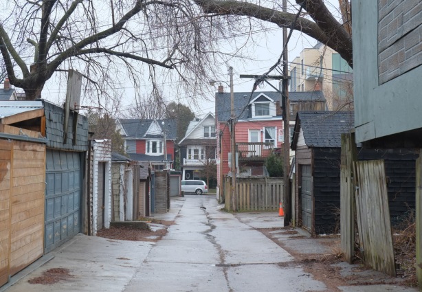 looking down an alley, with houses on the street at the end of the lane, garages, winter but no snow, fences, old trees, 