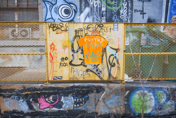 on upper and lower part of wall, fish graffiti, blue on top and pink below, in the middle is a yellowmetal railing with a yellow square covered with with graffiti including the outline of an orange t shirt 