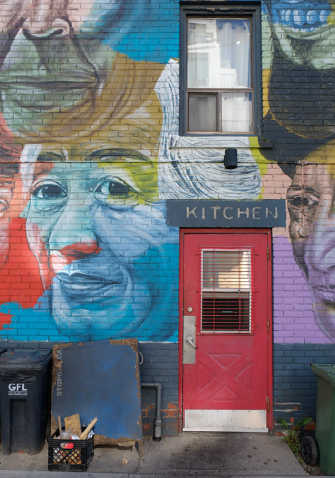 Troy Lovegates mural, faces by a red door with word kitchen over the top of it, blue face of a woman, also an upper storey window