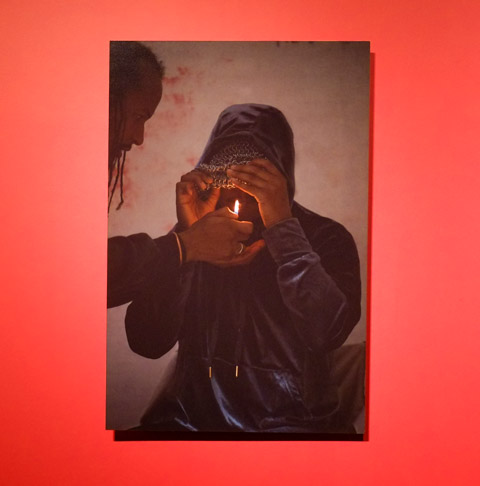 photograph on red wall, person helping to light a cigarette for man in a hoodie