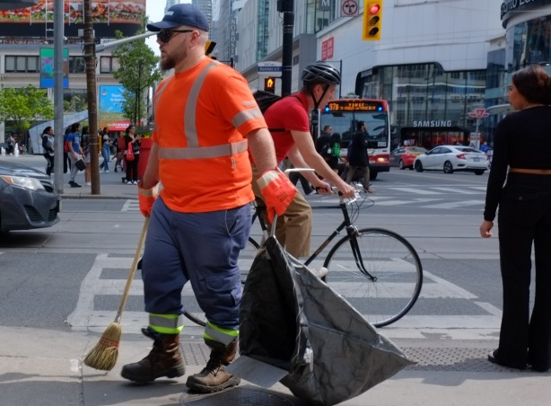 a garbageman in orange t shirt and large black bag walks past intersection of Yonge and Dundas, cyclist waiting for the traffic light to change