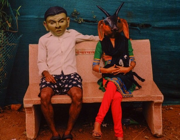 close up of photograph on a gallery wall, a couple sits on a bench, both wearing hindi masks. The man is in navy shorts with white stars on them and a long sleeved buttoned shirt, woman is in red leggings and an animal mask