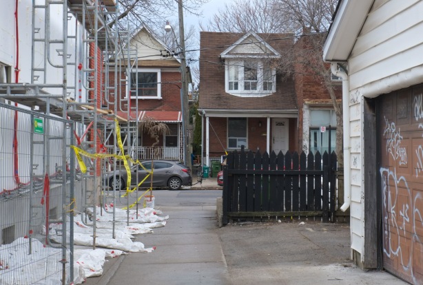 looking down a laneway to where it dead ends at a residential street with two storey brick houses, there is ablack picket fence in the alley, the house on the left in the alley is being renovated and has scaffolding covering the side
