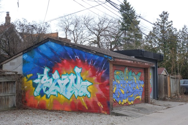 garage in an alley with street art on door and on side of building. on the side is white throwup on exploding red, yellow, and orange background