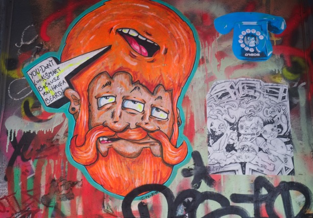 graffiti on a wall, pasteup of man with three eyes and a lot of orange hair and facial hair. Also a paste up by D76060 of a blue old fashioned rotary phone with an image of musician David Bowie in the center. 