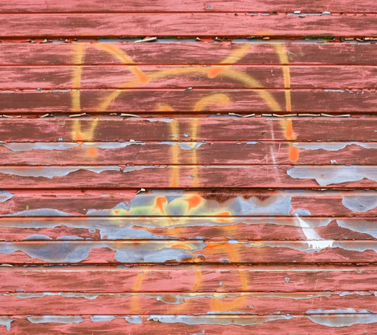 paint peeling on a reddish brown metal garage, old line drawing in yellow paint of a face, splotchy where bare metal can be seen 