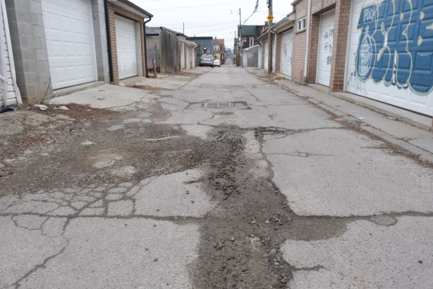 low view of alley showing mostly the pavement, cracked with dirt and wet spots, some garages along the sides
