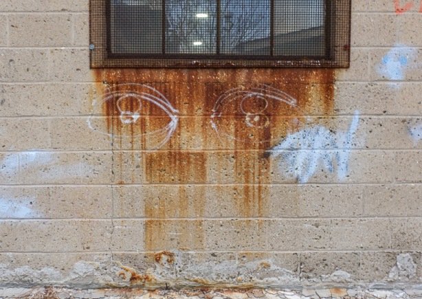 concrete wall under metal window frame, rust has stained the concrete, two white eyes have been drawn on the rusty part so now it looks like a face
