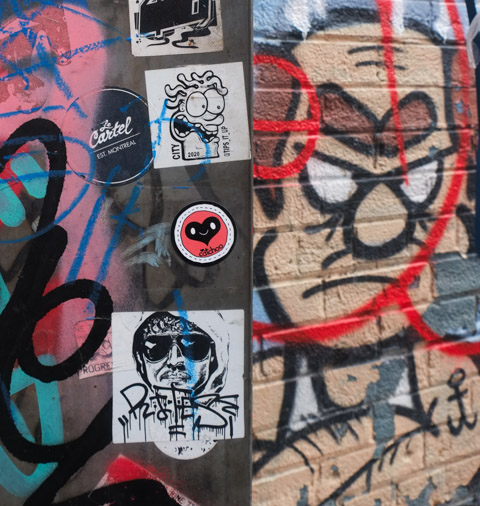 stickers, graffiti, on a pole, a painting of a scowling frowning man on the wall beside the pole 