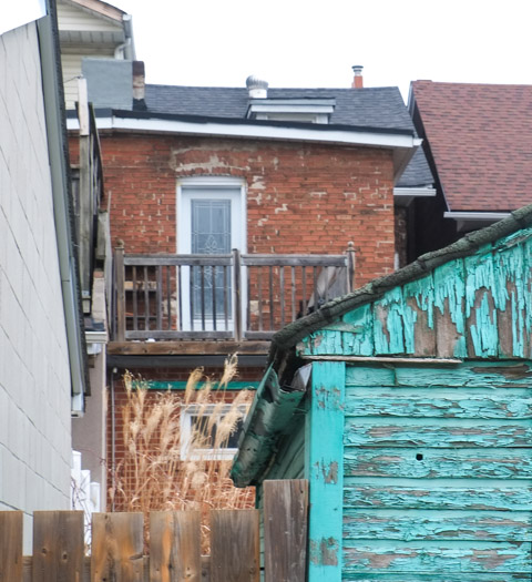 view from an alley, an old wood garage in peeling teal paint, two storey brick house with a small wood balcony on the upper level 
