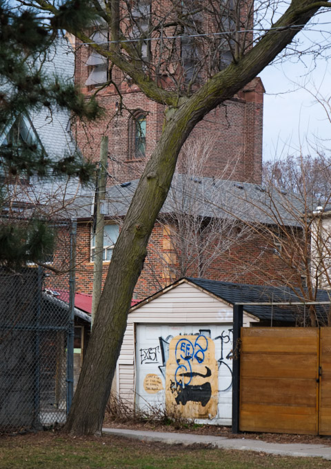 brick tower, perhaps steeple in the background, brick house in the middle, and a small garage with graffiti on door, large tree in the foreground