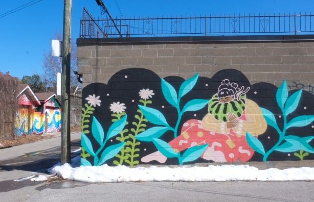 mural by caitlin ta in an alley, a woman is sitting, blue leaves on plants around her 