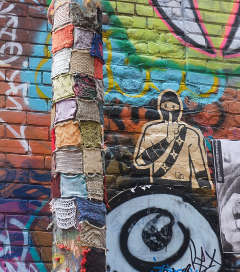 yarn squares, knit and or crocheted, and attached to a utility pole in alley, a large urban ninja squadron paste up is beside it 