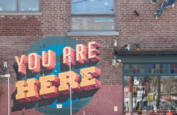 mural that says you are here in 3 d looking letters, on side of brown brick building 