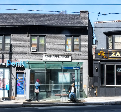 two people in a bus shelter, one standing, one sitting, in front of store called Atop Appliances which is an old grey building with a black shingle roof