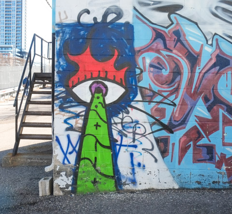 external metal staircase outside building beside a mural on the other wall, an eye, with red flames on the top, a green laser-like beam coming out of the eye
