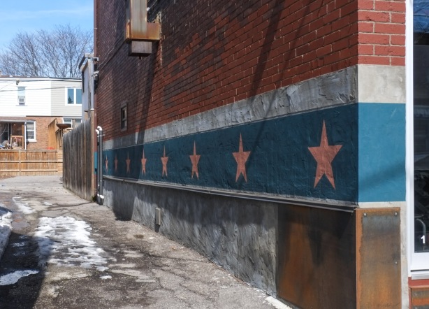 painted along the side of a red brick building, beside driveway, two stripes, a narrow white stripe on top and a wider blue stripe below.  On the blue, are eight pale orange stars