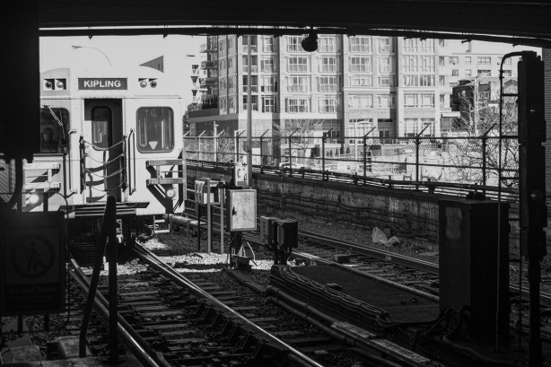 black and white photo of a TTC subway headed to Kipling, enters Keele station at section of track that is above ground, buildings in the background