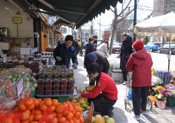 two young asian men working at a fruit and vegetable market, goods on sidewalk, some people passing by, a pile of orange in the foreground