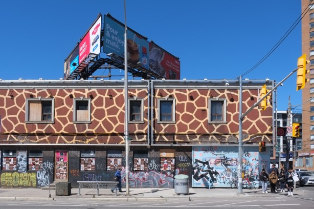 large billboard on top of two storey building with upper level painted like a giraffe in brown and yellow, bottom level covered with ads and graffiti