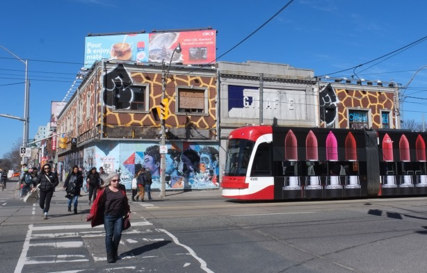northwest corner of Dundas and Bloor, people crossing with green light, TTC streetcar covered in ad for lipstick, giraffe building, 