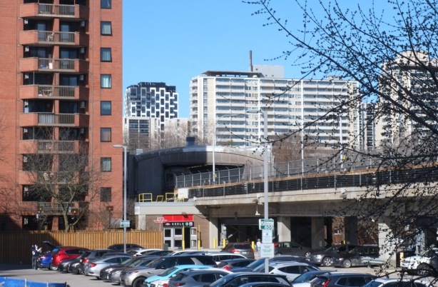 elevated subway tracks on east side of keele station, with parking lot below and pedestrian entrance, highrises in the background