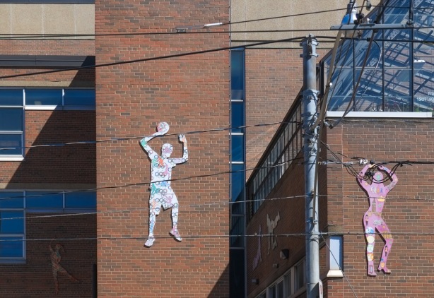 wood cutouts of people doing things, man holding a ball above his head, painted, mounted on exterior of red brick building, a school, 