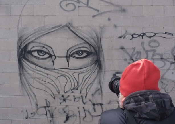 man in red wool hat taking a picture of anser eyes graffiti on grey wall