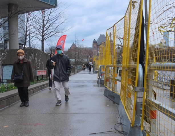 people walking in the rain on University ave where there is construction, yellow fence, Queens park building is in the background