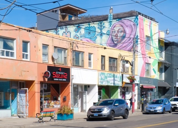 store fronts on gerrard street east, mural on the side of one building that is taller than the others, woman in a head scarf in mural, car parked on street, 