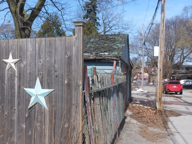 2 large stars as decorations, on old fence in an alley, crooked fence, car parked in alley, dead leaves on the ground, mossy roof on garage next door