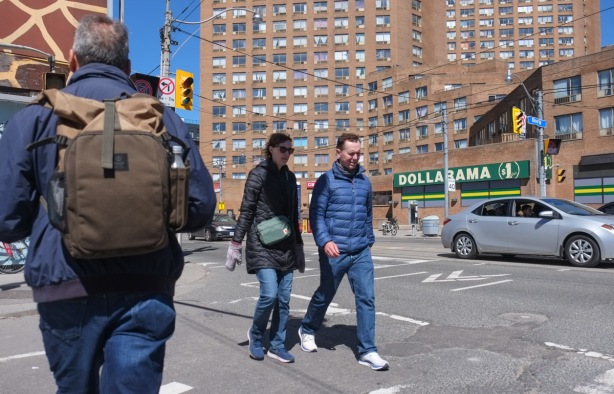 a couple walking together, crossing Bloor, with dollarama store and brick highrise apartment building behind them